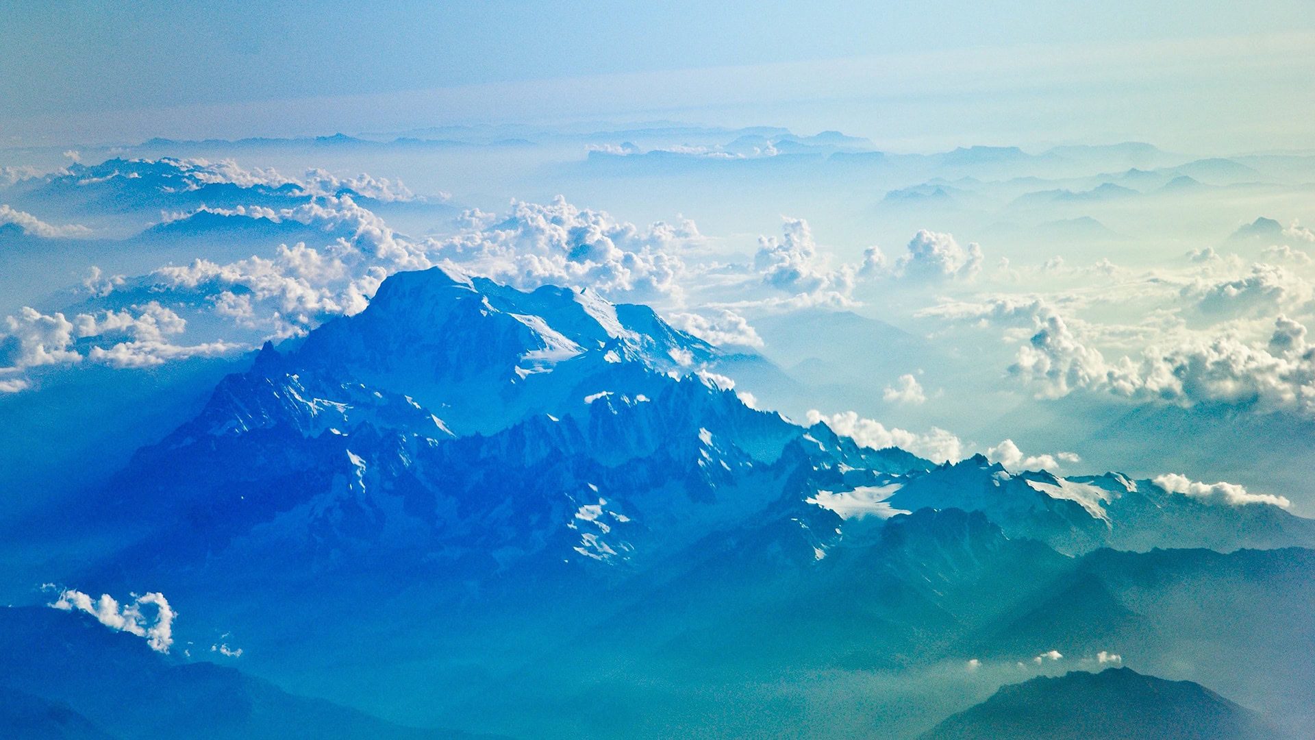 Aerial view of a large, snow-capped mountain surrounded by clouds under a blue sky.
