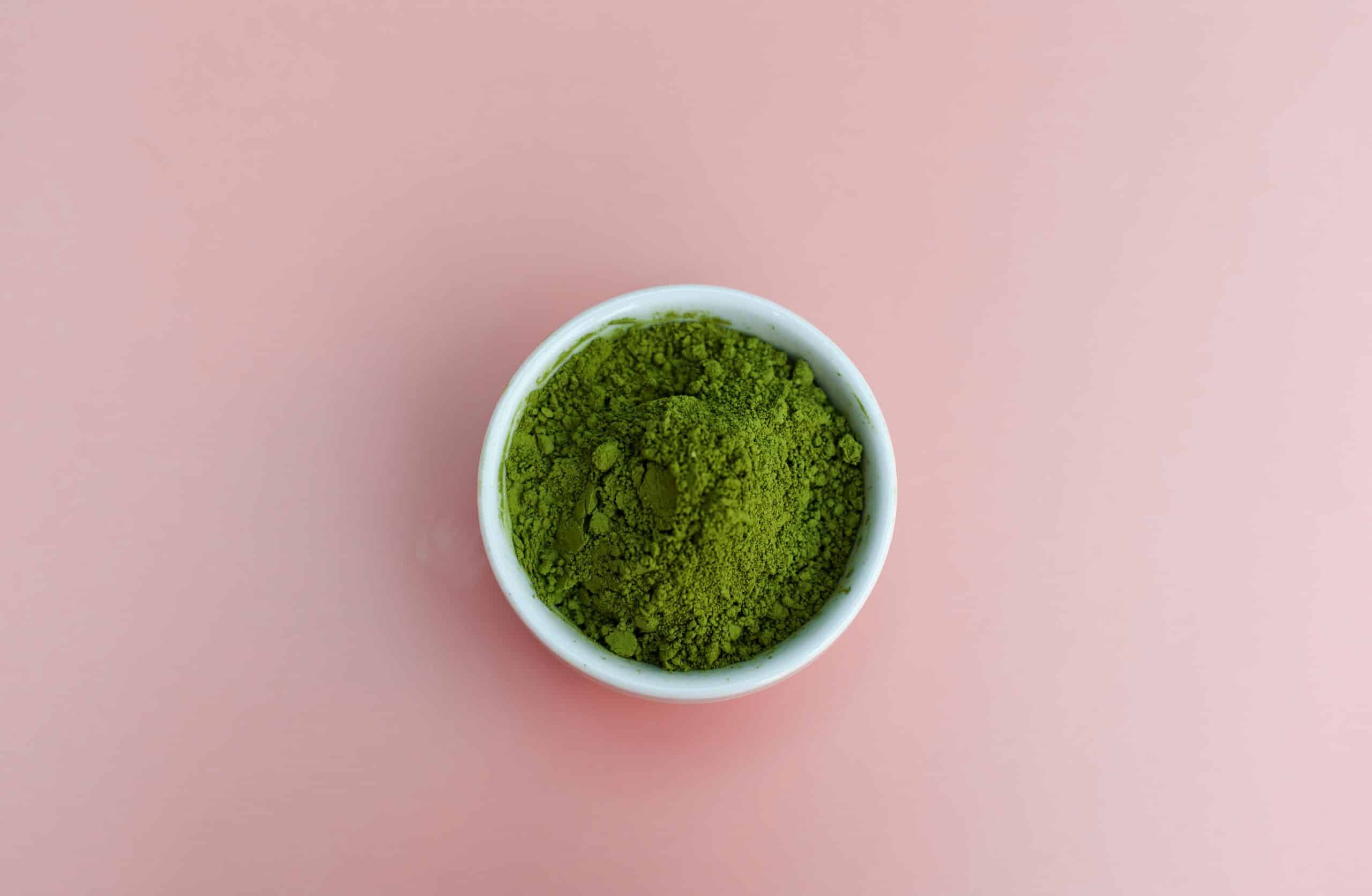 A small white bowl filled with green matcha powder sits against a plain pink background, highlighting the subtle difference between traditional tea and other botanicals like Mitragynine.